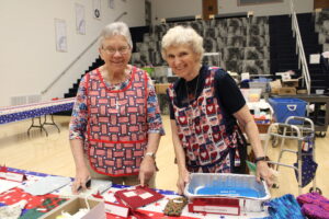 Two women in colorful aprons smile while working at a table filled with craft materials in a community space.