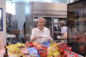 An elderly woman smiles while holding a plate in a snack area filled with various chip bags. Another person is seen in the background.